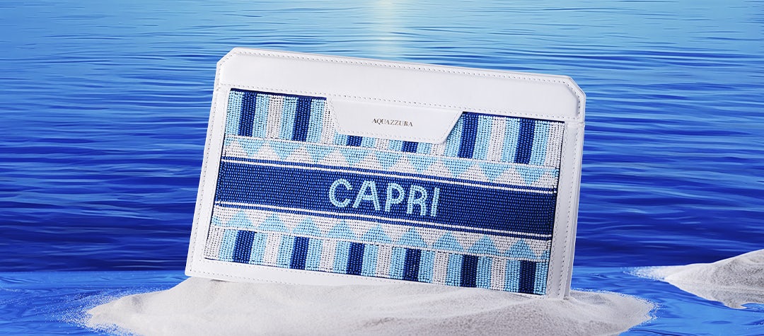 Named after the famous resort island, our white leather Capri Pochette features intricate beadwork in shades of blue and white on the front and a natural raffia panel on the back. The pochette in the image stands on a tiny stretch white sand that peaks out of the blue ocean.